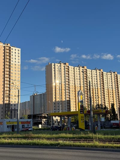 This vertically oriented image shows a street scene with tall, modern apartment buildings on the left and a gas station on the right. The sky is clear blue with a few wispy white clouds, and a sliver of the moon is visible. The sun is shining brightly, reflecting off the windows of the apartment buildings.
The apartment buildings are light beige and feature numerous windows and balconies. Power lines crisscross the foreground and extend towards the buildings.
The gas station appears to be part of a chain, with prominent yellow signage and red accents. Visible text on the gas station signs includes "РОСНЕФТЬ" (Rosneft), the name of a Russian oil company. Another sign shows fuel prices for different types of gasoline, such as "92", "95", "98", and "ДТ" (diesel fuel). A sign on a building within the gas station complex reads "МЫ РАДЫ ВАМ!" (We are glad to see you!).
The scene suggests it is daytime, likely late morning or afternoon, given the bright sunlight. The weather appears to be clear and sunny. The presence of a gas station and apartment buildings indicates an urban or suburban setting, consistent with the provided location context of Dnp Ryzhiki, Russia. There are no people clearly visible in the image.