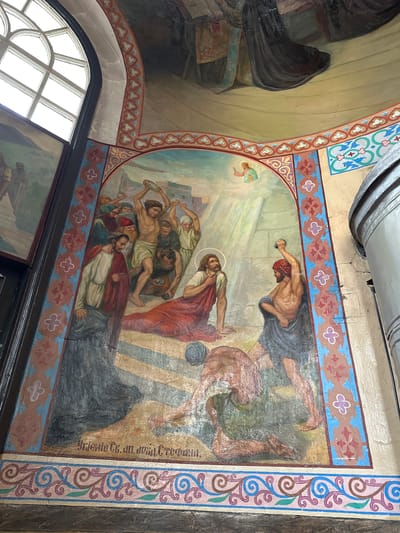 The image depicts a religious fresco on a wall, likely within a church or cathedral, possibly in Nogornyi, Russia. The fresco portrays a scene of persecution or martyrdom, with several figures in historical attire. In the center, a man with a halo, presumably a religious figure, kneels on a stone surface, facing a group of men. One man, to the right, is shown raising a stone or object above his head, seemingly about to strike the kneeling figure. Other figures around him are also depicted with raised arms and stones. A divine light beams down from the upper right, illuminating the scene. Decorative borders with floral and geometric patterns frame the fresco. The fresco shows signs of age and wear, with visible cracks and faded paint. A section of a large window with arched panes is visible in the upper left corner, suggesting the fresco is located in a space with natural light. The overall atmosphere is one of dramatic tension and suffering, common in religious narratives.