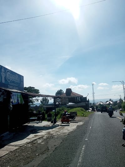 The image depicts a roadside scene under a clear, bright blue sky with scattered white clouds, illuminated by intense sun glare from the upper right. The location is Ka Thaung (Upper), Myanmar.
In the foreground, to the left, a dark blue commercial sign for "Crystalin" and "Toko Agung" is visible, affixed to a building with a corrugated metal awning. Underneath the awning, in deep shadow, two indistinct individuals are present on a concrete surface next to a dark cylindrical object. Hanging fabrics or clothes are visible below the awning. The ground beneath the awning and adjacent to the road is a mix of rough concrete and asphalt.
To the right of the concrete area, a paved two-lane asphalt road with a dashed white center line recedes into the distance, curving slightly right. An orange object, possibly a small vehicle or market stall component, is parked on the unpaved shoulder next to a black motorcycle. A white sign with the text "VILLA KANAN" is positioned nearby.
Further back, on an elevated grassy mound, a building with a distinctive brown, multi-tiered roof and a sign reading "SERVIS... OLI & MOTOR SPAREPART..." is visible. A tall, gray cellular tower stands behind this building. On the road, several vehicles are observed heading away from the viewer, including a dark SUV or car and at least two motorcycles, one with a rider wearing a light-colored helmet and jacket. In the midground and background, additional buildings with red-brown roofs are interspersed among green trees, set against a backdrop of distant hills or mountains. Overhead, power lines traverse the sky, and a street light pole is visible on the far right.