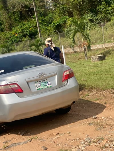 The scene depicts an outdoor environment in Odo Ona, Oyo State, Nigeria, under direct sunlight. A silver Toyota Camry sedan, approximately a 2007-2009 model, is positioned on an unpaved, gravelly dirt road, angled with its rear facing the viewer and slightly to the left. The vehicle's license plate, "OYO LUY-306AS NIGERIA," is fully legible. Partially obscured by the car's rear, a single individual is observed from behind, walking away from the camera. This person wears a dark-colored short-sleeved top, a light-colored headwrap with dark patterns tied at the back of the head, and carries a dark shoulder bag. To the right and behind the vehicle, a mown grassy area leads to a chain-link fence. Beyond the fence, dense green tropical foliage and trees cover a hillside. Within the grassy area, a small palm tree with reddish fruit clusters and a concrete block structure are visible. Distinct shadows are cast on the ground, indicating bright daylight.
