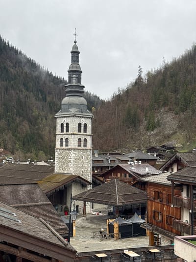 This is an eye-level, outdoor, vertical-format shot of La Clusaz, France, showcasing the church tower of Église Sainte-Foy de La Clusaz.

The shot is taken from a high vantage point, likely a building or elevated platform, providing a view that encapsulates the church tower, surrounding wooden buildings with steep roofs, and a backdrop of a wooded hillside shrouded in mist.

The church tower, constructed with light-colored stone, features arched windows and a multi-tiered spire topped with a cross. On the steeple face are circular clocks.

The surrounding buildings are predominantly constructed from wood, featuring traditional Alpine architecture with sloping roofs. The wooden roofs cascade down the buildings creating a geometric pattern.

The wooded hillside provides a natural backdrop to the urban landscape. The trees are dense, and a mist or fog hangs in the air, particularly at the base of the hill, adding a muted and atmospheric quality to the scene. The overall mood is subdued, reflecting a gray and overcast day.