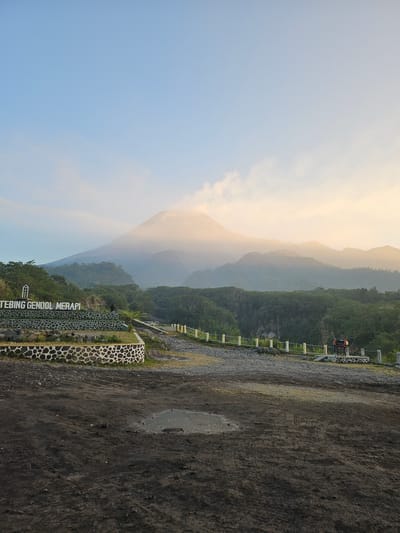 A wide-angle, eye-level shot captures a volcanic landscape at sunrise. The sky is a gradient of light blue and pale orange, with wisps of clouds. Dominating the background is a majestic volcano, its peak shrouded in mist, with layers of receding, green-covered hills fading into the distance. In the midground, a tiered stone wall and a sign that reads "TEBING GENDOL MERAPI" mark a scenic overlook. A few people are visible, one standing near the sign and two others in the distance by a red structure that appears to be a decorative art installation. The foreground consists of a dusty, dark-colored dirt ground with scattered small stones and a small puddle of water. The overall scene suggests a tranquil, natural setting in Cangkringan, Indonesia, known for its proximity to Mount Merapi.