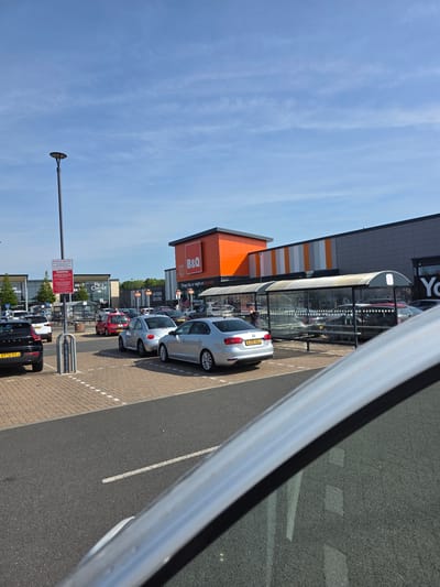 The image shows a view from inside a vehicle, looking out at a parking lot of a shopping area in Colchester, United Kingdom. Visible establishments include B&Q, Pets Corner, and an establishment labeled "Yo". Several cars are parked in designated spaces, including a silver car with license plate "BJ09 ABJ" and a black SUV with license plate "AY72 GVJ". A covered walkway runs along the front of some of the stores. There is a light pole and signs regarding parking restrictions. The sky is blue with wispy clouds.