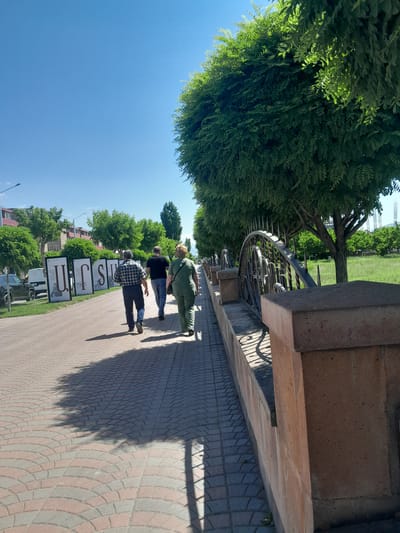On a bright, sunny day in Artashat, Armenia, three individuals walk along a paved pathway lined with trees. The pathway is constructed of interlocking red and grey pavers arranged in a fan pattern. To the left of the path, a row of large display boards featuring Armenian letters is visible, with vehicles parked behind them. The foreground is dominated by a stone wall and ornate metal fence. Further into the background, a grassy field with scattered trees and construction cranes can be seen under a clear blue sky.
