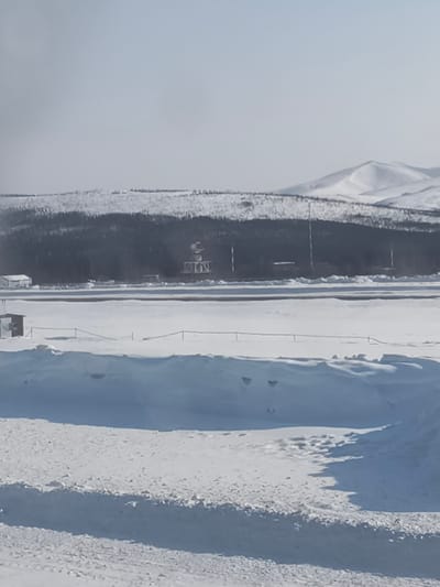 The scene depicts a snow-covered airfield environment in Sokol, Russia, under a bright, potentially hazy sky. The foreground is comprised of extensive snowdrifts and a textured snow-covered ground. Centrally, a dark, cleared strip, consistent with an airport runway or taxiway, extends horizontally across a broad, snow-laden expanse. To the left of this cleared strip, a small, dark, rectangular structure stands on the snow. In the midground, a dense line of dark evergreen trees forms a forest boundary behind the airfield. Within this tree line, a prominent lattice tower topped with a white spherical radome and multiple slender communication towers are visible. A white building, possibly an airport facility, is partially discernible on the far left within the tree line. The distant background features snow-capped mountains beneath a pale sky. No human figures or active vehicles are present in the observable area.