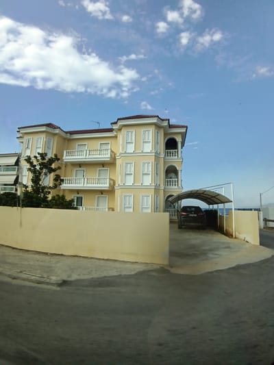 A yellow, multi-story building with white balconies and windows is viewed from a low angle on a sunny day. A dark SUV is parked under a white, arched carport. The building appears to be located in a coastal area, with the sea visible in the background beyond the carport. The scene is in the Municipal Unit of Sikyona, Greece.