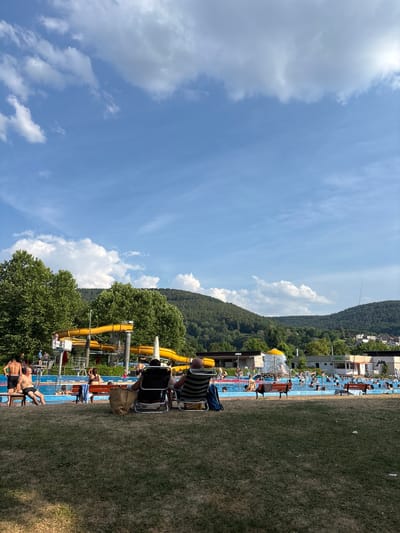 This media file captures a lively scene at an outdoor swimming pool complex. The primary subjects are numerous people enjoying a sunny day. The setting is a well-equipped water park, featuring multiple blue swimming pools and prominent yellow water slides winding through the landscape. A grassy area, likely a sunbathing lawn, occupies a significant portion of the foreground, with some people seated on chairs or loungers. Beyond the pools, a dense, green forest covers a hillside, with scattered buildings visible at the base and top of the slope, suggesting a town or village nestled against the natural environment.

The activity taking place is a day of recreation and leisure. People are seen swimming, relaxing by the pool, and preparing to use the slides. The atmosphere appears relaxed and enjoyable, typical of a summer day at a public pool. The weather is sunny with a bright blue sky dotted with white, fluffy clouds, indicating a pleasant day for outdoor activities. The time of day seems to be midday or early afternoon, given the strong sunlight and the active nature of the scene.

Notable details include the vibrant yellow of the water slides contrasting with the blue of the pools and the lush green of the surrounding nature. The presence of trees around the slides adds to the scenic beauty. Some people are dressed in swimwear, while others are casually attired for a day out. There are no discernible signs with visible text in the frame. The location context of Bürgstadt, Germany, suggests this is a local amenity for residents and visitors.