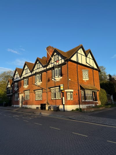 The image shows a multi-story Tudor-style building in Dacorum, United Kingdom. The building is constructed of red brick with white and black timber framing details on the gabled roof sections. Windows with diamond-shaped muntins are present. The image captures a section of the road with cobblestone pavement and yellow road markings. A bus stop is visible on the street next to a bench. The sky is clear blue. No people are visible.
