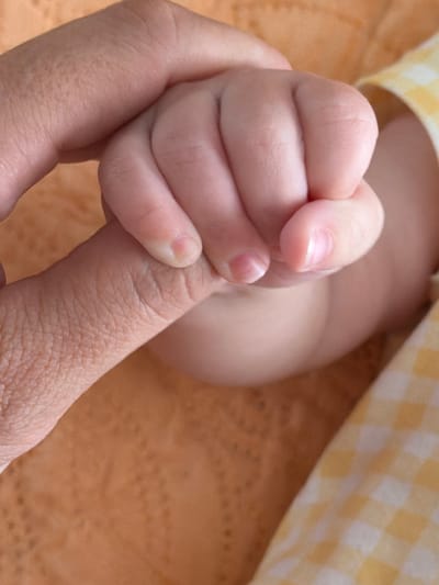 A close-up photograph shows a baby's hand gripping an adult's index finger. The baby's skin is smooth and light pink, while the adult's skin displays fine lines and wrinkles. The background is blurred, revealing a yellow and white checkered fabric and an orange textured surface. The location of this image is unknown and not specific to the city of Brasília, Brazil. The scene appears to be a moment of connection and support between a parent and child.