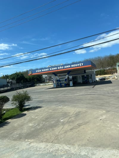 A gas station with a blue and orange canopy stands on a large concrete lot. Wires stretch across a clear blue sky to the left. Trees and other vegetation are visible behind the station and along the right edge of the lot. The ground is mostly gray and marked with lines and shadows. The scene is bright with sunlight.