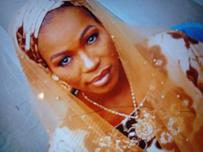 A woman wearing a traditional head covering, possibly a hijab or veil, and ornate jewelry poses for a portrait. The covering and attire appear to be in shades of beige, gold, and brown. She has a neutral expression, with her eyes gazing directly at the camera. The jewelry includes a necklace with a large pendant. Her makeup is done with defined eyebrows, eyeshadow, and lipstick. The image has a slightly soft focus, and the lighting is warm.