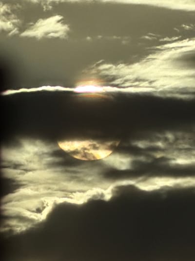 The image depicts a partially obscured sun seen through a cloudy sky. The sun, a glowing orb, is visible in the lower center of the frame, with its upper portion hidden behind dark clouds. Lighter, wispy clouds surround the sun and extend towards the top of the image, catching and diffusing the sunlight in a yellowish-white hue. The overall lighting suggests either sunrise or sunset. The scene is a natural landscape and does not contain any identifiable urban elements or people. The location of Krasnyi Krym, Russia, is not visually evident from the provided image.