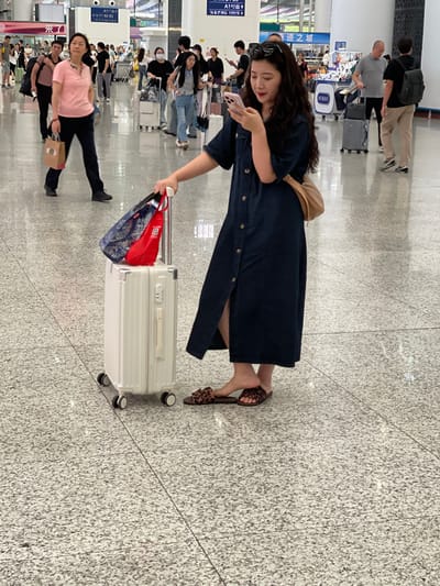 In a well-lit indoor public space, likely an airport or train station in Longhua District, China, a young woman with long, dark, wavy hair stands holding a white suitcase and a pink smartphone. She is wearing a dark denim dress and leopard-print slide sandals. Her phone is positioned in her right hand, and she appears to be looking at its screen. Two bags, one blue with a patterned design and one red, are attached to the handle of the suitcase. The suitcase has four wheels and a silver handle. In the background, numerous other people are visible, some walking with luggage, others standing. The floor is made of speckled gray tile. Signs in Chinese characters are visible on the walls in the distance.
