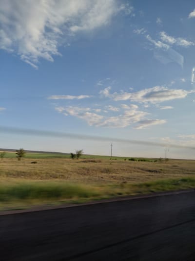 The image depicts a blurred, roadside view of a rural landscape under a blue sky with scattered white clouds. In the foreground, a dark asphalt road stretches horizontally across the bottom of the frame. Beyond the road, a field of dry, golden grass is visible, with a strip of green vegetation closer to the road. In the middle ground, a few sparse trees stand in the field, and in the background, there are rolling hills and what appear to be power lines. The overall impression is of a vast, open space. The image quality is low, with motion blur suggesting it was taken from a moving vehicle. There are no people or specific structures indicative of the city of Молодёжный, Kazakhstan. The scene is natural and unpopulated.