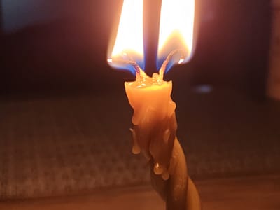A close-up, eye-level shot shows the top of a twisted, beeswax candle with two wicks burning in a dimly lit environment. The flames are bright orange and white, with a hint of blue at their bases, indicating intense heat. Melted wax drips down the sides of the candle, forming irregular shapes. The background is dark and out of focus, with subtle variations in color that suggest a textured surface. There are no people visible in the image. The scene is devoid of any identifiable landmarks or context that would place it specifically in Bugulma, Russia.