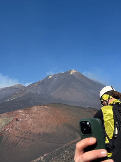 A person is captured from behind, holding a dark green-cased smartphone in their right hand, pointed towards an active volcanic landscape. The individual wears a white helmet, a bright yellow head covering or bandana, a yellow vest over a dark long-sleeved shirt, and a black backpack with a silver bottle attached. Dreadlocks or braided hair extend from beneath the helmet. The smartphone's triple camera array is visible. The scene is set on the slopes of Mount Etna, near Nicolosi, Italy, under a clear blue sky. The prominent volcanic cone in the background displays dark grey and reddish-brown terrain with visible yellowish deposits around its peak, indicating volcanic activity. White plumes of smoke or steam emanate from multiple vents on the volcano. In the midground, winding dirt paths are visible across the reddish-brown volcanic soil, with several small groups of distant hikers traversing them. The foreground consists of dark, rugged volcanic rock and soil.