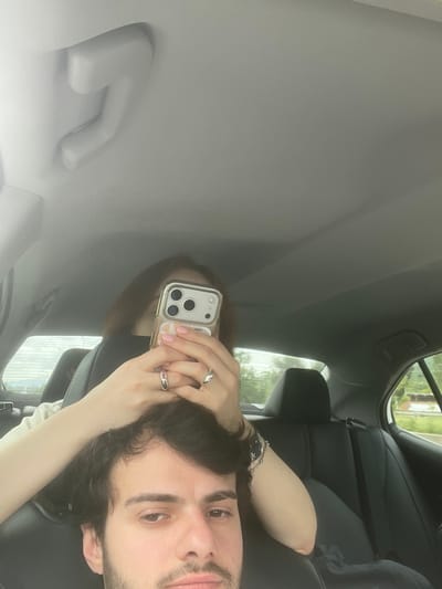 The image depicts a man sitting in a car, presumably in Changilari, Georgia. He is holding a phone in front of his face, with his left hand on the phone. The car's interior is visible, including the headliner, grab handle, seats, and a glimpse out the window. The lighting suggests daylight.