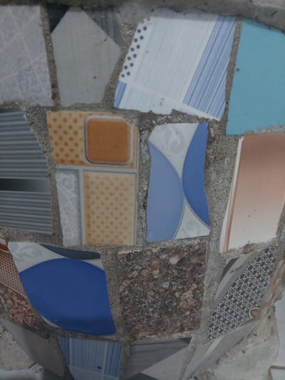 A close-up, low-angle, vertical shot shows a mosaic made of broken ceramic tiles adhered to a concrete surface. The mosaic consists of various fragmented tiles in different colors and patterns, including white and gray geometric designs, blue and white stripes, solid blue, orange with polka dots, and speckled brown and white tiles. Some tiles have a glossy finish, while others appear matte. The texture of the concrete grout is visible between the tile pieces. The overall impression is of a decorative art piece, possibly on an outdoor structure or furniture. This mosaic is located in the City of Pematang Siantar, Indonesia.