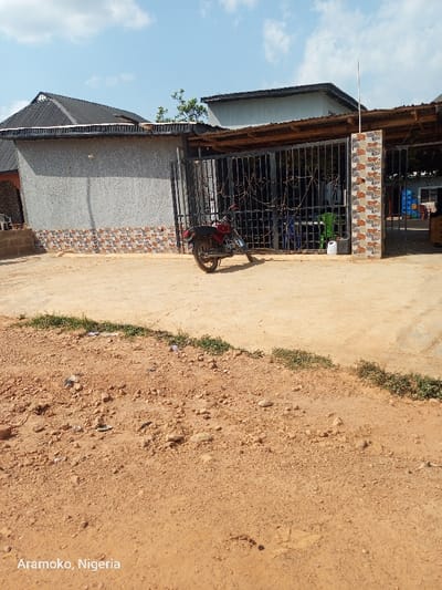 The image shows the front of a building with a grey wall, tiled halfway up with brown and white tiles. The building is surrounded by a black metal gate. There is a black motorcycle with a red seat and a black fender parked in the dirt, facing the gate, with the front tire of the motorcycle turned to the left. There is a large metal door on the right side of the image which is open, revealing a space with chairs and tables, and a small shop behind the door. The gate is closed. There is a grassy area in the foreground, and the image is taken from a slight angle. There are no people in the image, and the sky is clear and blue. The scene is in Aramoko, Nigeria at the coordinates 7.836, 4.476.