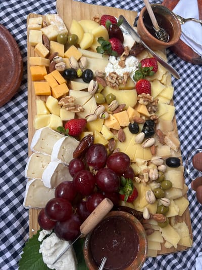 A wooden board is laden with a variety of cheeses, including brie, cheddar, and other hard and soft varieties. The cheeses are complemented by fresh strawberries, red grapes, dates, olives, pistachios, almonds, and walnuts. Two small clay pots hold honey and jam. The spread sits on a black and white checkered tablecloth.