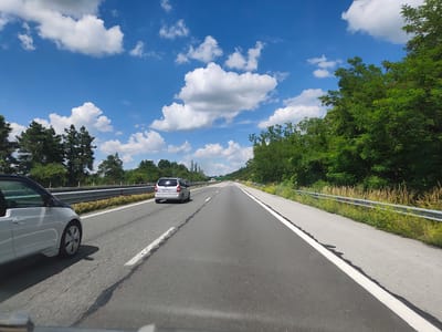 This media file captures a scenic view from a car traveling on a highway, likely during the day. The scene is set against a backdrop of a bright blue sky dotted with fluffy white clouds, suggesting fair weather. Lush green trees line the right side of the highway, adding to the natural beauty of the location.
The highway itself is paved and appears to be a multi-lane road, with clear white lane markings indicating the direction of travel. In the foreground, a white car with a black roof is partially visible on the left, suggesting the perspective is from inside another vehicle. Further down the road, a silver hatchback is in the left lane, followed by other vehicles in the distance. A metal guardrail runs along the side of the highway, separating the road from the vegetation.
The overall impression is one of a pleasant journey through a rural or semi-rural area. There are no people explicitly visible, but the presence of cars indicates ongoing travel. The time of day is clearly daytime, given the brightness and the shadows cast. The location, identified as Ravnishte, Bulgaria, provides a geographical context. There is no discernible text within the image that provides additional information. The mood conveyed is tranquil and serene, typical of a road trip on a sunny day.