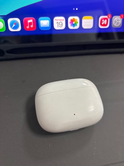 A white, oval-shaped charging case for wireless earbuds is positioned on a dark gray, textured surface. The case appears slightly dusty, with a visible seam running across its top and a small dark circular indicator light on its front. Behind the case, a portion of a screen displays a row of app icons on a blue background. These icons include what appears to be Safari, Music, Mail, Calendar, Photos, Notes, and possibly Yandex and Settings. The scene is lit from above, casting a shadow of the earbud case onto the surface. The location is specified as Imeni Morozova, Russia, but there are no visual cues in the image to confirm this geographical information.