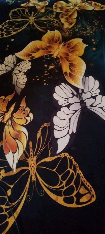 The image is a close-up, overhead shot of a black fabric with a repeating pattern of stylized butterflies. The butterflies are depicted in various shades of yellow, orange, and white, with some outlined in gold. Some butterflies have intricate wing patterns, while others are more abstract. The fabric appears to be a textile material, possibly for clothing or home decor. There is no indication of any specific location in Boraldai, Kazakhstan, or any people, actions, or interactions within the image. The image solely focuses on the decorative pattern of the fabric.