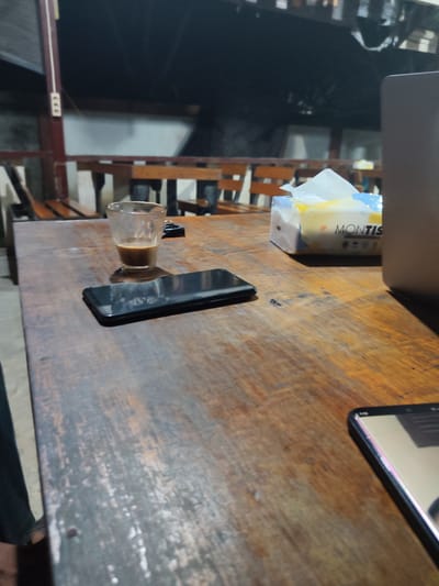 A wooden table at a cafe in Keudematangkuli, Indonesia, is shown. On the table, there is a partially full glass of coffee, a smartphone, and a box of Montis tissues. A laptop is also visible on the right side of the image. The cafe appears to be outdoors, with wooden seating in the background. The lighting suggests it is evening or night.