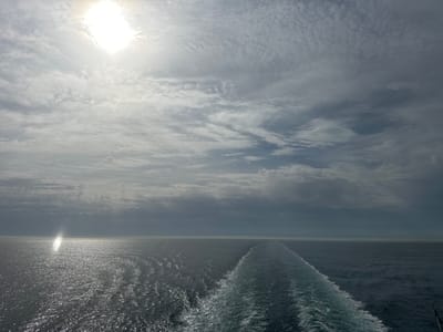 The image depicts a seascape under a cloudy sky. The sun is visible through the clouds, casting a bright glare. The water surface is ruffled, reflecting light. A ship's wake stretches into the distance, suggesting movement across the water. No specific city in France is identifiable from this image.