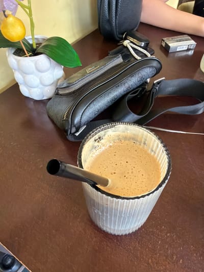 A close-up shot captures a brown wooden table holding a glass of frothy brown beverage with a black straw, a black fanny pack with a patterned design and zippers, a car key with a decorative fob, a pack of cigarettes, and a white pot containing a plant with green leaves and a yellow decorative fruit. In the background, a portion of a person's arm, light-skinned and wearing a bracelet, is visible, suggesting they are seated at the table. The scene is set within a cafe or restaurant in Sapareva Banya, Bulgaria, indicated by the presence of these common cafe items and the table's surface texture.