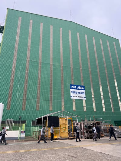 The image depicts a large, green, corrugated metal industrial building, likely a shipyard building, located in Geoje-si, South Korea. A sign on the building reads "2C4 AREA" in English, and in Korean, "선행의장 전용작업장" which translates to "Outfitting workshop for the front". Below this, another Korean phrase reads "해양공사(주)선행의장부", potentially indicating the company name "Ocean Construction Co., Ltd. Outfitting Division".
In the foreground, several individuals, likely workers, are walking on an asphalt path. They are dressed in casual work attire, including pants and jackets, and several are carrying backpacks. To the left of the building, there is a yellow structure with a hazard symbol indicating radiation. Next to it, a metal fence encloses a small area with signage. Various metal scaffolding and materials are stacked to the right of the yellow structure. The sky is overcast.