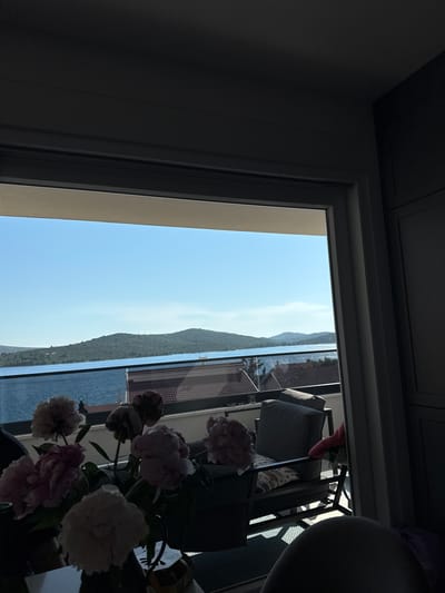 A view from inside a room looks out through a large window to a balcony and then to a seascape under a clear blue sky. On the balcony, there is outdoor furniture including chairs with cushions. In the foreground, inside the room, there is a vase of pink and white peonies. The background shows a body of water, likely the Adriatic Sea, with islands and hills in the distance. The scene appears to be in the city of Šibenik, Croatia, possibly overlooking the Dalmatian coast. The lighting suggests it is daytime.