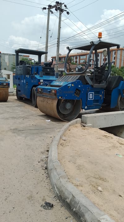 The image shows a construction site in Effurun, Nigeria. Three blue Hamm brand road rollers are parked on an asphalt surface adjacent to a newly constructed concrete curb and a section of sandy fill. The rollers are positioned in a line, with the closest roller prominently displaying the model number "C2865 B". Electrical poles with multiple wires run vertically in the background. A building with "SIGNATIO" visible on its side is partially seen behind the road rollers. The sky is overcast with light-colored clouds. The scene appears to be a road construction or repair project within an urban environment. There are no visible people in the image.
