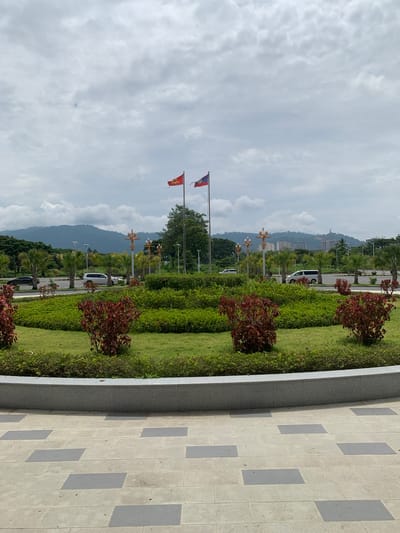 The image captures a wide-angle view of a landscaped area in the city of Golden Triangle Special Economic Zone, Laos. In the foreground, a circular paved area made of light grey and dark grey rectangular tiles leads to a raised, curved granite border. Beyond the border, a lush green lawn is dotted with several small shrubs with dark red foliage. Behind the lawn, a circular arrangement of neatly trimmed green bushes forms the centerpiece. In the mid-ground, a paved road runs horizontally across the image, with several vehicles, including a black sedan, a white SUV, and a white minivan, driving or parked alongside it. Palm trees and decorative lampposts line the road. Two flags are visible on flagpoles in the distance; one is red with a yellow symbol, and the other is red with a blue stripe and a white circle in the center. In the background, a range of low mountains and a city skyline with several buildings, including some under construction, are visible under a cloudy sky. The overall scene suggests a well-maintained public space or entrance area within a developed urban environment.