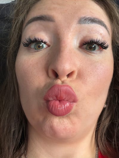 A close-up, head-on shot shows a woman with long, brown hair, her face framed by dark brown hair. She is wearing dramatic false eyelashes and her eyes are a striking green-brown. Her lips are plump and pursed, coated in a glossy pink lipstick. The woman has a light dusting of freckles across her cheeks and nose. The background is dark and out of focus, with a hint of what appears to be dark upholstery to the left. The lighting is soft, highlighting the contours of her face and the texture of her skin. The woman appears to be making a kissing gesture. There is no indication of the location in the city of Старые Туймазы, Russia.