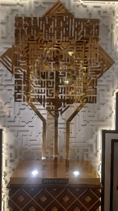 A detailed, full-shot image captures an elaborate golden metallic artwork displayed indoors in Glantengan, Indonesia. The central subject is a large, multi-dimensional sculpture composed of intricate, angular Arabic calligraphy, specifically forming the text of "Ayat Kursy" in a Kufic-like script. This golden sculpture is supported by multiple angled, polished gold-colored struts that descend to a rectangular golden pedestal.
Embedded within the golden pedestal are three distinct circular spotlights, emitting bright white light upwards to illuminate the artwork. A dark rectangular plaque is affixed to the front of this golden pedestal, bearing the English text "AYAT KURSY" in white capital letters. The golden pedestal rests upon a substantial wooden base or cabinet. This lower wooden section features a decorative marquetry pattern of alternating light and dark wood squares and diamonds, each intersection adorned with a small, circular golden inlay.
The background consists of a light-colored wall covered in a repeating, embossed geometric pattern, characteristic of Islamic architectural design. No individuals are present in the scene. The lighting is focused on the artwork, with ambient illumination on the patterned wall.