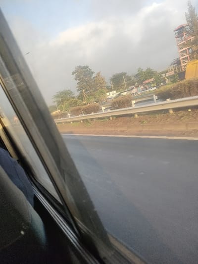 The image depicts an oblique exterior view captured from within a vehicle, showing a paved road and the surrounding environment in Theta ward, Kenya.

In the immediate foreground on the left, elements of the vehicle's interior are visible, including a dark window frame and dark fabric, possibly a seat or clothing. The primary subject is a multi-lane asphalt roadway, dark grey in color, extending from the midground into the distance. A solid white line delineates the right edge of the visible road segment.

Parallel to the road, a white metal guardrail is present, separating the road from an earthen strip. Beyond this strip, a row of low, bushy green and brown shrubs forms a median or roadside barrier. In the background, numerous trees with varied foliage densities and heights line what appears to be another roadway or area with commercial structures. Distant buildings are partially obscured by the trees.

To the far right of the frame, a distinctive, multi-story structure with an exposed, reddish-brown skeletal frame, possibly a tower, stands prominently against the skyline. Several vehicles, including at least one white car, are discernible on a road or parking area beyond the shrubbery. The sky above is partly cloudy, showing patches of light blue and scattered white-grey clouds, indicating daylight conditions. No individuals are clearly visible.