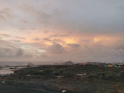 An elevated, panoramic view at dusk or dawn captures a coastal and semi-arid landscape within Granadilla de Abona, Spain. The upper two-thirds of the image depict an overcast sky, transitioning from diffused orange-pink hues near the horizon to a duller grey-blue overhead. The sea forms a calm, grey horizontal expanse in the distance, meeting the land horizon.
The midground features varied terrain and structures. To the left, large white-roofed buildings, possibly greenhouses or industrial coverings, are visible close to the shoreline, accompanied by sparse, dry vegetation and a prominent, bare, skeletal tree. In the central midground, a distinct cone-shaped hill, identifiable as Montaña Roja, rises in the distance. A tall metal lattice tower stands near its base. Industrial buildings with various roof types are scattered across the landscape. To the right, several brightly colored shipping containers (red, blue, green, yellow) are arranged linearly, indicating a storage or commercial zone. Artificial lighting from streetlamps is discernible in this area. The foreground consists of dark, uneven ground covered with sparse, low-lying scrub and volcanic rock. No individuals or active human interventions are visible in the scene.