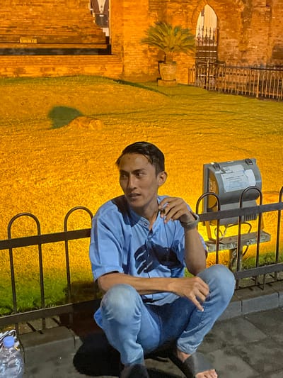 A male subject is observed squatting on a dark paved surface at night in Kauman, Indonesia. He is positioned in the foreground, facing forward with his head turned towards the right, looking upwards. The subject wears a light blue collared short-sleeve shirt and light blue jeans, with dark sandals partially visible on his feet. A lit cigarette is held between the index and middle fingers of his right hand, which also bears a dark wristband. His left hand rests on his left knee. A clear plastic water bottle is visible on the ground to his lower left. Behind him is a low, black metal fence. The background is brightly illuminated with a warm, golden-orange light, showcasing a patch of green grass directly behind the fence, and further back, a large brick structure. This structure features an arched doorway or gate on the right, containing a dark metal gate, and tiered brickwork or steps on the left. A potted plant is positioned in the grass area in front of the brick structure. To the right, a grey utility box or device is visible on a stand at the edge of the grassy area.