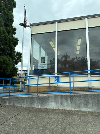The image shows the exterior of a post office in Beaverton, Oregon, United States of America. A flagpole with an American flag is visible to the left of the building. The building has large windows with a reflection of trees and overhead lights. A sign on the window indicates that U.S. passports can be applied for inside. A blue railing lines a ramp leading up to the building, and a blue handicap symbol is attached to the railing. The environment appears overcast.