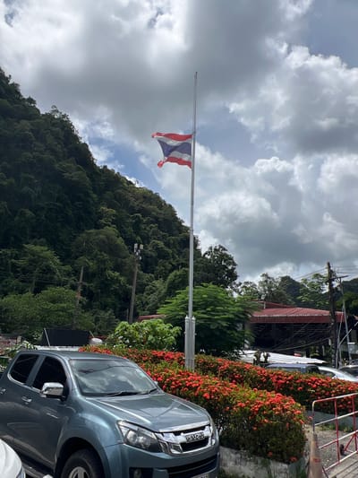The image shows a flagpole with a Thai flag flying. The flag is partially obscured by clouds and is positioned against a backdrop of a lush, green, mountainous landscape. In the foreground, a dark-colored pickup truck is parked. The truck has the word "ISUZU" visible on its front grille. To the right of the truck, there are bushes with bright red flowers and a red and white striped barrier. Electrical wires and poles are also visible, suggesting an inhabited area within a natural setting. The sky is overcast with heavy clouds, indicating potentially cloudy or rainy weather. The scene appears to be set in a tropical or subtropical region, consistent with the location context of Tambon Wang Prachan, Thailand. There are no people visible in the image, and the overall mood is one of natural scenery with signs of human presence.