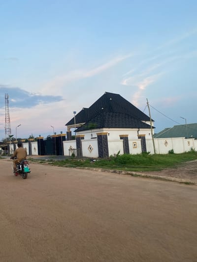 In the city of Uffah, Nigeria, a man rides a motorcycle on an unpaved light brown dirt road, moving away from the viewer towards the left. He is dressed in a light brown tunic and red footwear. The motorcycle features a green rear mudflap and dual exhaust pipes. To the right of the road, a two-story house with a distinct black shingled roof, multiple gables, and white exterior walls is visible. The property is secured by a white perimeter fence accented with dark grey panels and gold detailing, including an ornate main gate. A small solar panel is mounted on a pole near the roofline of this house. Further right and partially obscured, another property features a green roof and a white fence with diamond-shaped gold inserts. In the distant left background, a tall, metallic lattice communication tower stands prominently. A weathered wooden utility pole with a black cable is positioned on the right side of the main house. The sky above is light blue with sparse, thin clouds, gradually shifting to a pale orange-pink at the horizon. Patches of green grass and weeds grow alongside the dirt road.