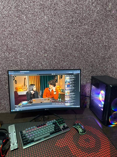 The image displays a computer setup in a room with dark, textured carpeting. The scene is captured from a slightly elevated perspective, looking down at a desk area.
On the left side of the frame, a black desktop computer tower is visible. It features a transparent side panel, revealing internal components, most notably several fans that are illuminated with vibrant, multi-colored RGB LED lighting, creating a rainbow effect.
Dominating the center of the image is a large, black computer monitor. It is displaying a video, likely from YouTube, given the visible user interface elements. The video features two individuals:
* In the upper portion of the video frame, a man with curly dark hair and an orange long-sleeved top is seen. He is seated at a desk with a microphone in front of him, looking slightly downwards or to the side. The background behind him includes a dark green wall and some objects on a shelf.
* In the lower portion of the video frame, a woman with long, straight dark hair and a black top is visible. She is also seated at a desk with a microphone. Behind her, an acoustic guitar hangs on a wall, and several circular objects (possibly vinyl records) are displayed on a shelf above what appears to be a dark red couch or chair.
The monitor's interface shows a list of video thumbnails or a playlist above the main video. A dark grey progress bar is visible at the bottom of the video player. In the bottom left corner of the monitor, several browser tabs are open, with one clearly displaying the YouTube logo. Partially legible text in the bottom right of the video player includes "MUSIC MONSTERS" followed by "L.A.B - ...". The browser's address bar shows "youtube.com/watch...".
To the right of the monitor, a black mousepad with a repeating pattern of red-orange dots covers a significant portion of the desk surface. On this mousepad, a gaming mouse with green LED accents and a distinct design is positioned towards the upper right. Below the mouse, a full-sized mechanical keyboard with multi-colored RGB backlighting is also present. Further down and partially off-frame, a silver, foldable portable gaming device (resembling a Nintendo Switch Lite) and some black cables or headphones are visible on the desk.
The floor throughout the visible area is covered with a dark grey or charcoal-colored shaggy carpet, providing a soft and textured surface.