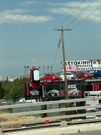 The image depicts a car dealership in Polichni Municipal Unit, Greece, featuring cars displayed on multiple levels and a large billboard. A red car is suspended upside down, seemingly as a promotional display, on a raised platform. Below, several cars are parked on a showroom roof, including red, white, and blue vehicles. A prominent billboard with Greek text, possibly advertising car sales and financing, is visible in the background. A wooden utility pole with electrical wires runs diagonally across the frame, obscuring parts of the scene. The environment includes buildings and trees in the distance, under a partly cloudy sky. A metal railing and a concrete barrier are in the foreground, suggesting the image was taken from a road or elevated walkway.