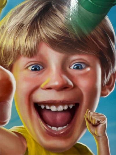 The image is a close-up, digitally rendered portrait of a young boy with blond hair and bright blue eyes, depicted with an exaggerated, wide-mouthed smile. He is wearing a yellow shirt and a green hat or accessory. The background is a solid blue sky. The boy's facial features, particularly his eyes, teeth, and rosy cheeks, are rendered with high detail and a vibrant, slightly unreal quality. His right hand is clenched and raised near his face, while his left hand is partially visible on the left side of the frame. There are no discernible elements within the image that would indicate a specific location in Keila, Estonia.