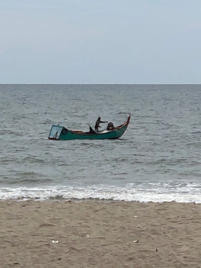 The image depicts a person in a small, green boat on a body of water. The boat is narrow and appears to be a traditional fishing vessel. The person, seen from a distance, is standing or crouching within the boat. The water is a light gray color with ripples, suggesting it is the sea or a large lake. A wave is breaking on the right side of the image, indicating a shoreline is present. The shoreline itself is a sandy beach with a rough, textured appearance. The sky is a pale blue-gray, hinting at an overcast or hazy day. The image is oriented vertically, with the top of the boat pointing towards the bottom of the frame. The overall scene suggests a tranquil, possibly early morning or late afternoon setting, given the soft lighting. The location is identified as Keudekruenggeukueh, Indonesia. There is no visible text in the image.