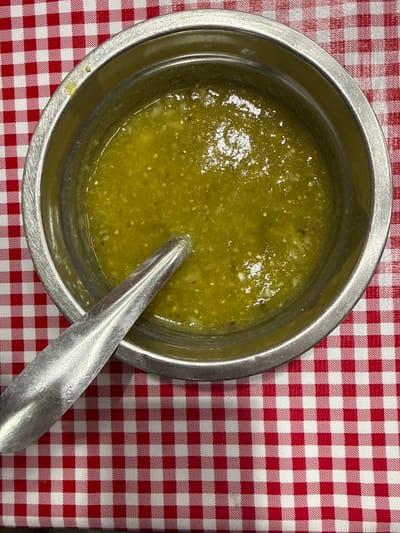 A top-down, static shot presents a close-up view of a stainless steel bowl filled with a viscous, chunky green salsa. The salsa, consistent with salsa verde, exhibits a vibrant light green color with visible lighter flecks of pulp and darker specks, likely seeds and finely minced herbs/vegetables. A polished silver-colored metal spoon rests partially submerged within the salsa, its handle extending diagonally from the lower-left corner of the bowl towards the center. The bowl and its contents are situated on a flat surface covered by a red and white gingham-patterned tablecloth. No individuals or active interactions are present. This scene is located in Nezahualcóyotl, Mexico.