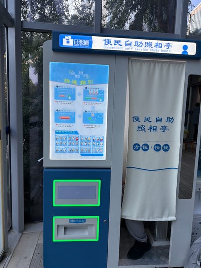 A self-service photo booth, blue and white, is depicted outdoors in Jianye District, China. The booth features a top illuminated sign displaying "证照通 (ZHENG ZHAO TONG)" and "Convenient self service photo booth." The left panel presents a large, illuminated screen with instructions titled "自拍自助快拍指引" (Self-service quick photo guide) detailing four steps, above a grid of small profile picture icons. Below this screen, the blue base incorporates two rectangular sections; the upper one is a blank display, and the lower one is a print output slot labeled "和冲印出口." To the right, a white curtain with "便民自助照相亭" (Convenient Self-Service Photo Booth) and "方便·快捷" (Convenient·Fast) indicates the photo-taking compartment. The lower legs and black-shoed feet of an individual in grey trousers are visible inside the booth, positioned for use. The background, visible through reflective glass panels, shows green foliage, suggesting an outdoor or semi-outdoor public setting.