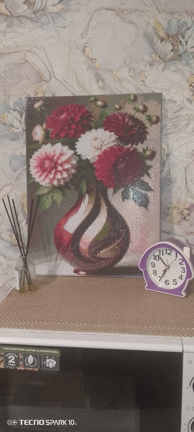 This is a medium shot, eye-level photograph featuring a still life arrangement on a shelf. The background is a textured wallpaper with a floral pattern in muted tones of beige, grey, and light blue.

The primary subject is a framed diamond painting depicting a bouquet of flowers in a vase. The flowers are rendered in shades of deep red, pink, and white, with green leaves. The vase is a deep, glossy red with swirling patterns of black and white. The diamond painting is positioned centrally in the frame, leaning against the wallpaper.

To the left of the painting, a small glass bottle filled with diffuser reeds stands upright. The reeds are dark brown and extend above the bottle.

To the right of the painting, a small, purple house-shaped alarm clock is visible. The clock has a white face with black numerals and hands, indicating the time is approximately 10:06.

The objects are placed on a textured, light brown mat or placemat that covers the surface of a white appliance, which appears to be a microwave. The front of the appliance is black and displays several logos and text, including "TECNO SPARK 10c." The top edge of the appliance is white.

The overall lighting is soft and diffused, suggesting an indoor setting. There are no people or animals present in the image.

The image does not provide any information about the location being in the city of Srednii, Russia.