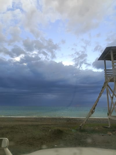 The image shows a beach scene in Gulrypsh, Abkhazia. A lifeguard tower stands on the right, constructed of light-colored wood. The ladder leans against the tower. The sky is overcast with dark storm clouds. A faint rainbow can be seen through the clouds toward the center of the image over the sea. The sea is a pale greenish-blue. The beach is composed of a dark sand strip and a strip of pebbles. In the foreground is a patch of green grass, partially obscured by the bottom edge of a vehicle window with the side mirror partially visible on the left.